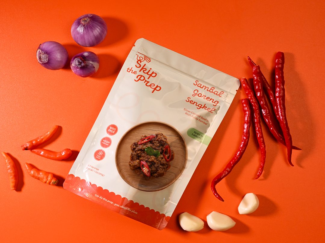 A pouch of Skip The Prep Sambal Goreng Sengkel surrounded by fresh chilies, shallots and garlic